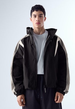 Mladý muž, ktorý má na sebe čierno-béžovú bundu s kapucňou a svetlosivé tričko, stojaci pred jednoduchým svetlým pozadím.