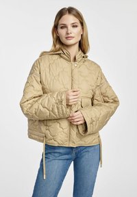 Beżowa pikowana kurtka z kapturem, z przodem zapinanym na napy, bocznymi kieszeniami i regulowanym ściągaczem u dołu, noszona na niebieskich dżinsach.