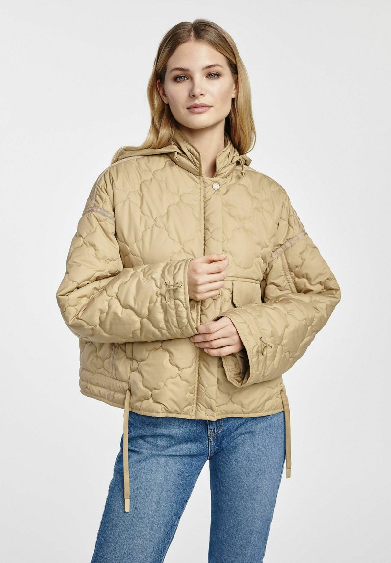 Beżowa pikowana kurtka z kapturem, z przodem zapinanym na napy, bocznymi kieszeniami i regulowanym ściągaczem u dołu, noszona na niebieskich dżinsach.