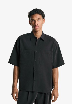 Black short-sleeve shirt made from a smooth fabric, featuring two chest pockets and a button-down front. Straight cut, relaxed fit.