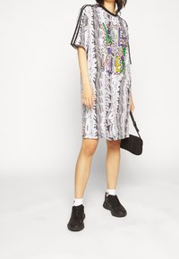 Vestido gráfico grande y gris con texto vibrante "LOVE UNITE", rayas verticales y mangas cortas; combinado con zapatillas negras y un bolso.