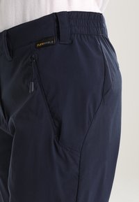 Close-up of navy blue pants with zipper pocket and FLEXSHIELD fabric label on the waistband against a plain background.