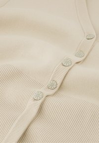 Cream-colored knitted cardigan with ribbed texture and five floral-patterned buttons along the front opening.