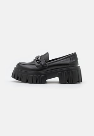 Mocassin à plateforme en cuir noir avec semelle crantée épaisse et chaîne décorative sur le dessus, vu de côté sur fond blanc.