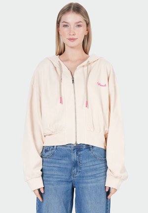 Jeune femme portant un sweat à capuche rose clair zippé avec le texte « Miracle » et un jean bleu, debout devant un fond blanc uni.