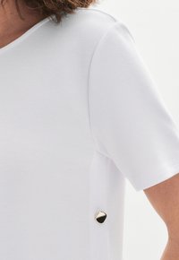 Camisa blanca de manga corta con una textura suave que presenta un detalle acentuado en dorado con un diseño geométrico en el lateral. Diseño clásico de cuello redondo.