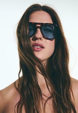 Młoda kobieta o długich brązowych włosach, nosząca duże ciemne okulary przeciwsłoneczne, widoczne nagie ramiona, neutralne jasne tło.