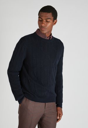 Jersey de punto trenzado azul marino con cuello redondo, tejido texturizado y puños de canalé, combinado con pantalones de cuadros marrones.