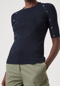 Námořnicky modrý krátký top s krátkými rukávy z žebrovaného úpletu se stříbrnými cvočky podél ramene. Má přiléhavý střih a měkkou texturu.