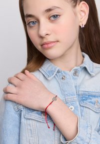 Młoda dziewczyna z niebieskimi oczami, nosząca kolczyki w kształcie motyla i jasną dżinsową kurtkę, pokazująca bransoletkę z czerwonego sznurka z zawieszką w kształcie serca.