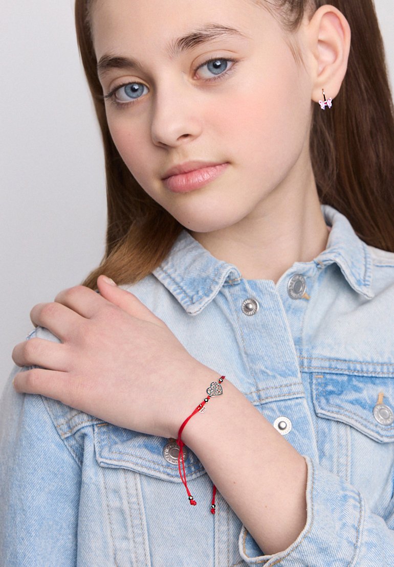 Młoda dziewczyna z niebieskimi oczami, nosząca kolczyki w kształcie motyla i jasną dżinsową kurtkę, pokazująca bransoletkę z czerwonego sznurka z zawieszką w kształcie serca.