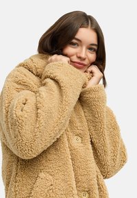 Veste en polaire beige texturée avec un col haut et de grands boutons. Son design confortable présente une coupe décontractée et des coutures bien visibles.