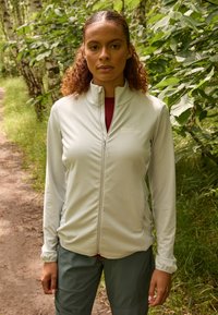 Lichtgrijze zip-up jack gemaakt van zacht materiaal, voorzien van een staande kraag en voorzakken. Draag het over een rode top. De achtergrond is weelderig groen.