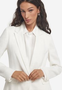 Vit figursydd kavaj i slät tyg, med slag med hack, framfickor och knappstängning, bärs över en ljusfärgad skjorta.