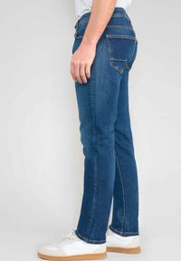 Jean en denim bleu foncé avec une coupe slim, un design à cinq poches et des accents brodés. Associé à des baskets blanches avec une semelle beige.