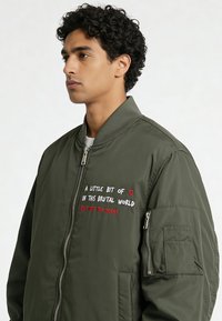 Olijfgroene bomberjack met geborduurde tekst aan de voorkant, ritssluiting en geribbelde manchetten. Voorzien van zijzakken en een ritszak op de arm.