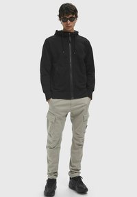 Felpa nera con zip e cappuccio regolabile, abbinata a pantaloni cargo beige chiaro con più tasche e scarpe da ginnastica nere.