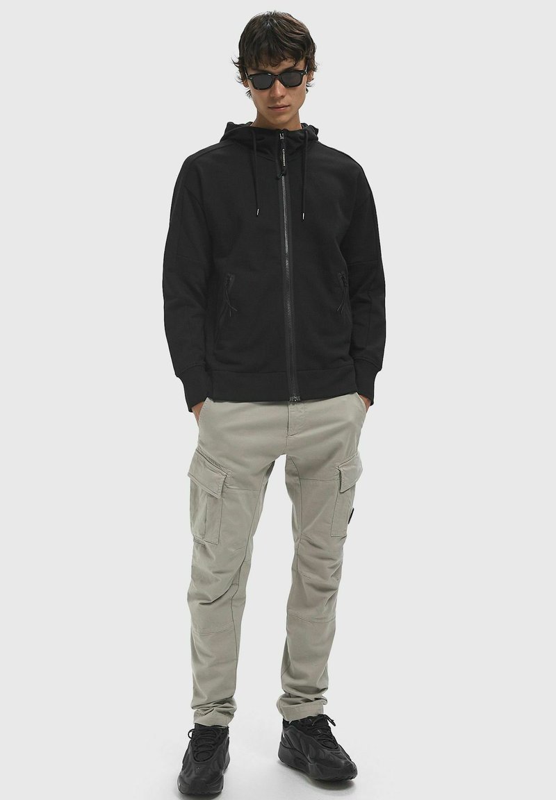 Felpa nera con zip e cappuccio regolabile, abbinata a pantaloni cargo beige chiaro con più tasche e scarpe da ginnastica nere.