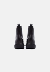 Ζευγάρι μαύρων δερμάτινων μποτάκια αστραγάλου με θηλιές για έλξη, ορατή ραφή και χοντρές σόλες, εμφανιζόμενα από πίσω σε λευκό φόντο.