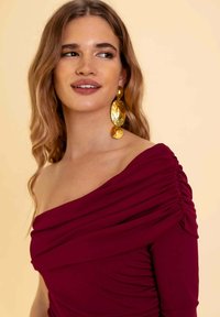 Vestido burdeos sin mangas con detalle fruncido, caída en los hombros y textura suave. Grandes pendientes dorados llamativos complementan el look.