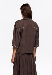 Brązowa, lekka lniana koszula o luźnym kroju, z rękawami 3/4 i kontrastowym przeszyciem wzdłuż tylnego szwu. Posiada prosty dół oraz wiązania.