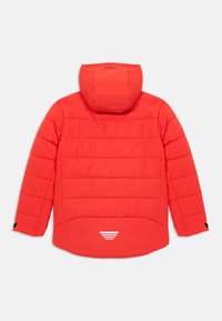 Casaco acolchoado vermelho com capuz, apresentando costuras horizontais, uma bainha curva e detalhes refletivos nas costas. Textura suave, sem fechos visíveis.