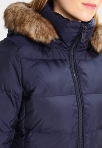 Donna che indossa un giubbotto imbottito blu navy con zip frontale e cappuccio finto pelliccia marrone, accorciato alle spalle.