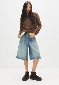 Tricou maro, cămașă galbenă dedesubt, șorturi din denim bleu deschis și botine maro până la gleznă. Design simplu, cu o croială relaxată și material texturat.