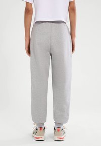 Osoba stojąca tyłem, ubrana w jasnoszare spodnie dresowe, białą skróconą koszulkę, białe skarpetki oraz beżowe buty sportowe z koralowymi akcentami.