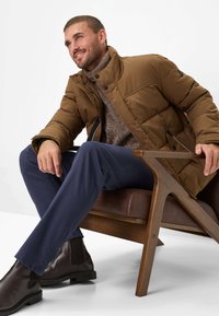 Strukturierte braune Pufferjacke mit Stehkragen, marineblaue Hose und dunkelbraune Chelsea-Boots, vor einem minimalistischen Hintergrund.