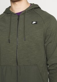 Olivově zelená mikina na zip s texturovanou horní částí, hladkou spodní částí, kapucí, stahovací šňůrkou a malým černým logem Nike na hrudi.