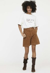 Shorts en velours côtelé marron avec des plis sur le devant et des poches latérales, associés à un t-shirt graphique blanc ornée d'un design imprimé. Bottines marron à la cheville.