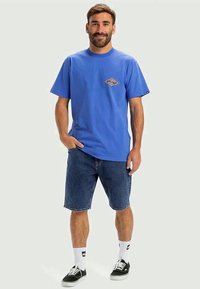 Μπλε βαμβακερό T-shirt με κεντρικό σχέδιο μάτι, συνδυασμένο με τζιν σορτς. Το μοντέλο φορά λευκές κάλτσες και μαύρα sneakers. Σχεδίαση τακτικής εφαρμογής.