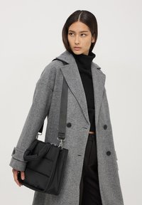 Borsa nera a sacco con superficie testurizzata e manici piatti. Il modello indossa un cappotto grigio in lana e un dolcevita nero.