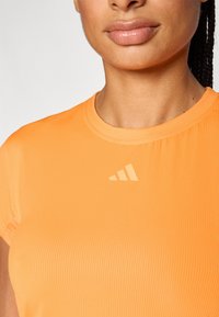 Tricou sport portocaliu cu textură răsucită, având un guler rotund și un logo bej în partea superioară stângă.