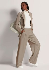 Beige colber met lange mouwen, gecombineerd met hoog-waist beige broek. Witte turtleneck en witte sneakers. Grote lichtgekleurde tas.