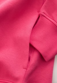 Nahaufnahme eines leuchtend pinken Sweatshirts mit geripptem Bündchen und Seitentasche mit detaillierter Naht.