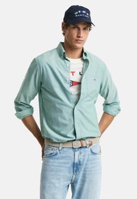 Lichtgroen overhemdd met opgerolde mouwen, gedragen over een wit graphic T-shirt. Lichtblauwe jeans en een marineblauwe pet met kleurrijke logo's.