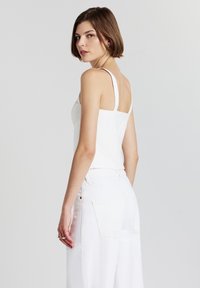 Giovane donna con capelli corti castani, che indossa una canotta bianca senza maniche e pantaloni bianchi, che si gira leggermente per guardare sopra la spalla.