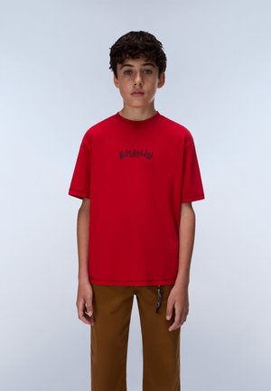 Garçon aux cheveux bouclés et foncés, portant une chemise rouge à manches courtes et un pantalon marron, debout devant un fond clair uni.