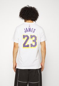 Hvid t-shirt med korte ærmer, der har lilla og gule "JAMES" og "23" trykt på bagsiden, samt NBA-logo ved halsen.