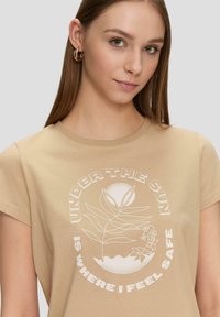 Beige katoenen t-shirt met korte mouwen. Bevat een witte grafische afbeelding van planten en de tekst "ONDER DE ZON VOEL IK ME VEILIG."