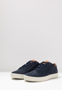 Mörkblå lädersneakers med vita gummisulor, bruna läderdetaljer på tungan och matchande snörning. Sömnadsdetaljer synliga.