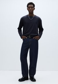 Temno modra majica Henley z dolgimi rokavi in gumbi na prednjem delu, kombinirana z ohlapnimi temnimi jeansi iz trapera in črnimi škornji s pasnim poudarkom.