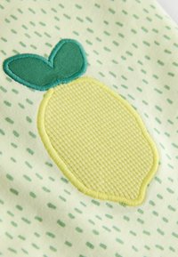 Citronformad gul brodyr med gröna blad på en ljusgul tygbakgrund med ett prickigt mönster i gröna nyanser.