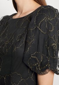 Robe noire avec des manches flottantes transparentes ornées de motifs floraux brodés en or, mettant en valeur la texture et le détail complexe.