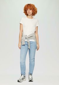 T-shirt blanc à manches courtes, jean bleu clair, sweatshirt gris noué à la taille et chaussures de sport argentées. Tissu lisse et coupe ajustée.