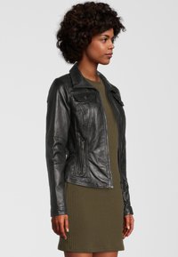 Zwarte leren jas met een getailleerd ontwerp, voorzien van een kraag, twee voorzakken en een ritsluiting. Gecombineerd met een groen ribgebreid jurkje.