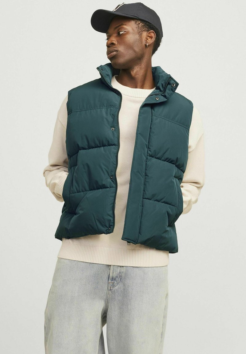 Donkergroene puffer vest met hoge kraag, ritssluiting en drukknoopjes. Draag het over een crèmekleurige trui, met gewatteerde voering en zijzakken.