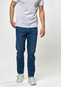 Kék farmernadrág egyenes szabással, klasszikus öt zsebes kialakítással, enyhe kopott hatással és tiszta, felhajtott szegéllyel. Fehér pólóval viselve.